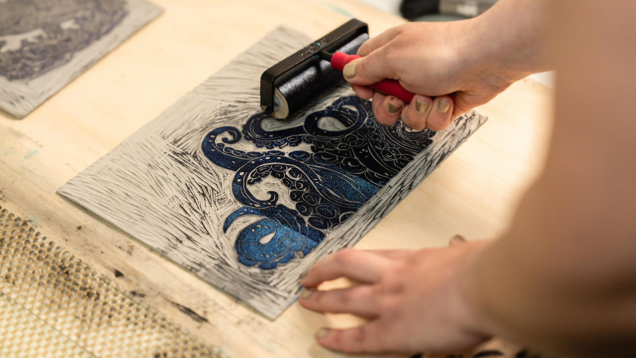 A person uses a brayer to ink a linocut print depicting swirling kraken tentacles. The scene conveys artistic focus and creativity on a wooden surface.