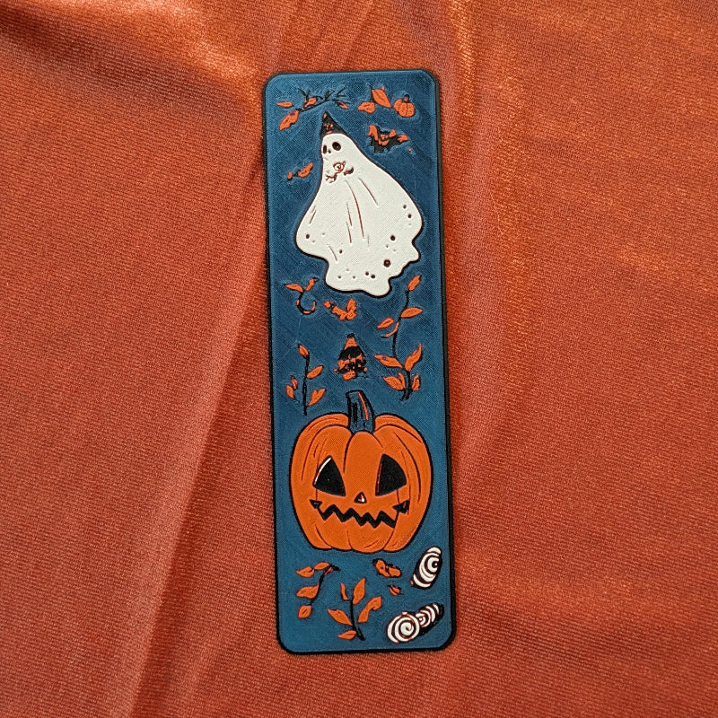 Textured 3D printed ghost and pumpkin bookmark with autumn leaves on deep blue background