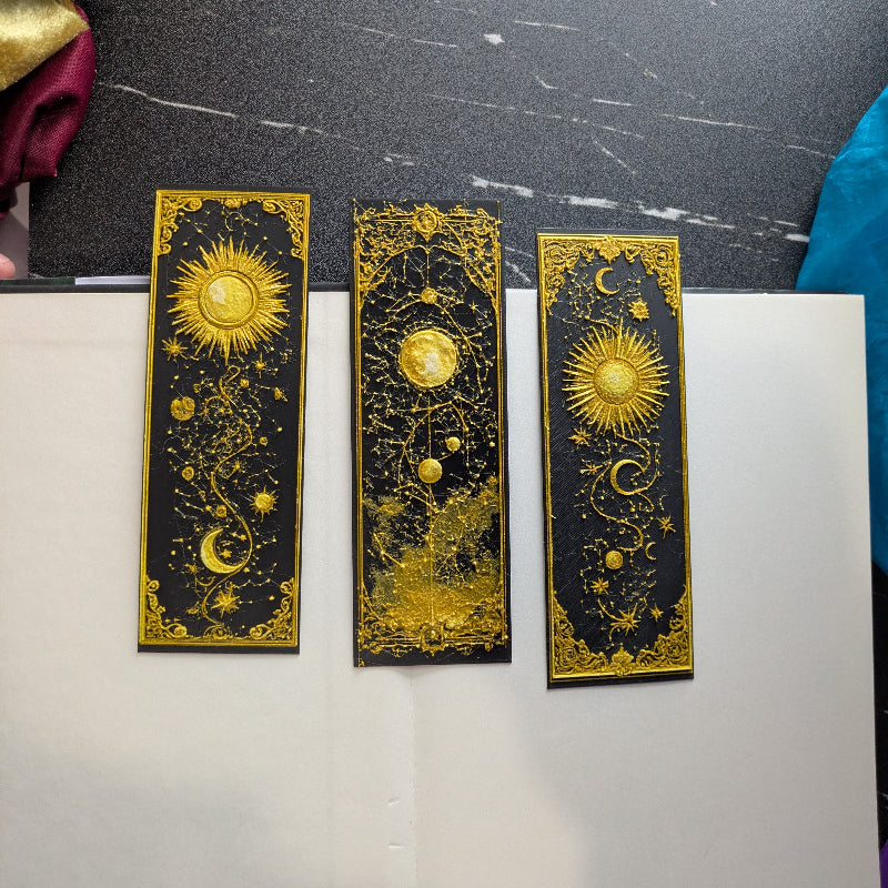 Three gilded celestial bookmarks displayed together in black and gold with sun, moon, stars, and ornate borders