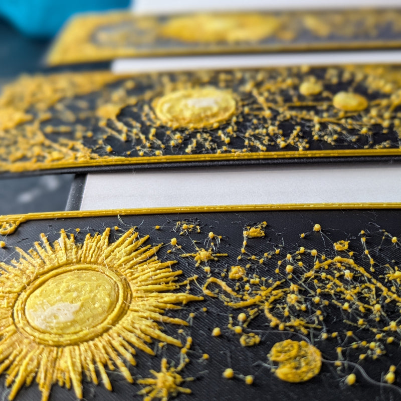 Close-up of three gilded celestial bookmarks showing textured gold celestial artwork and ornate decorative borders