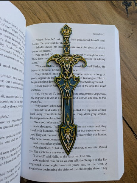 Gilded sword-shaped bookmark with gold and blue ornate detailing resting on an open book, inspired by medieval and fantasy designs.