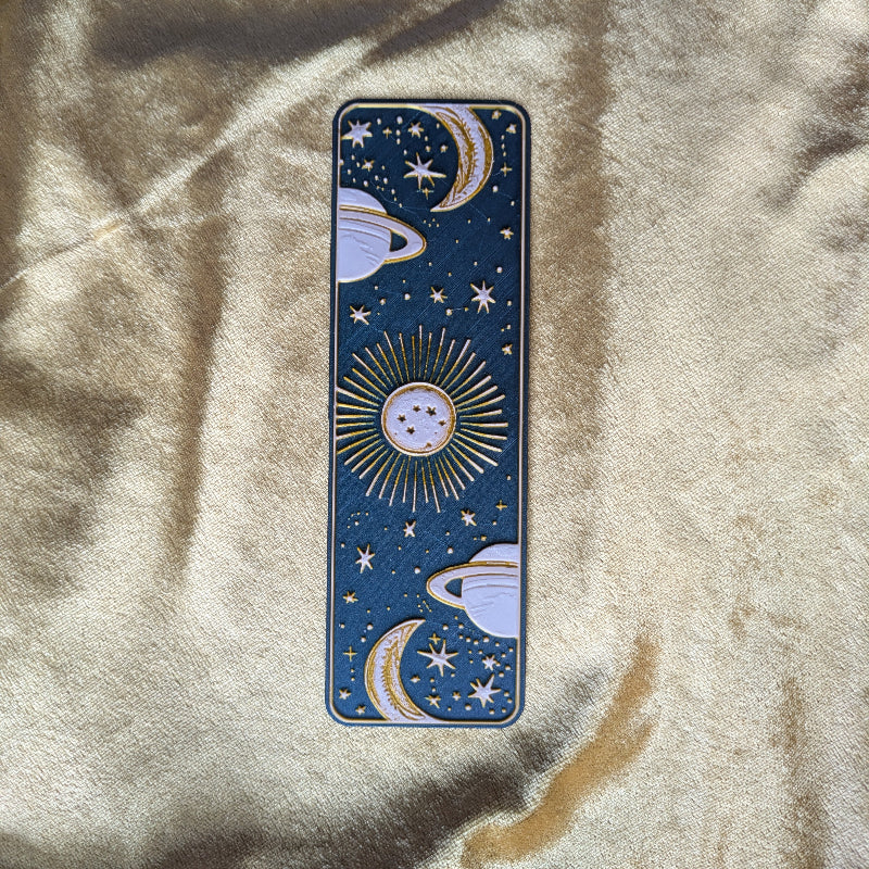 Gold embossed Saturn and crescent moon celestial bookmark on deep teal background