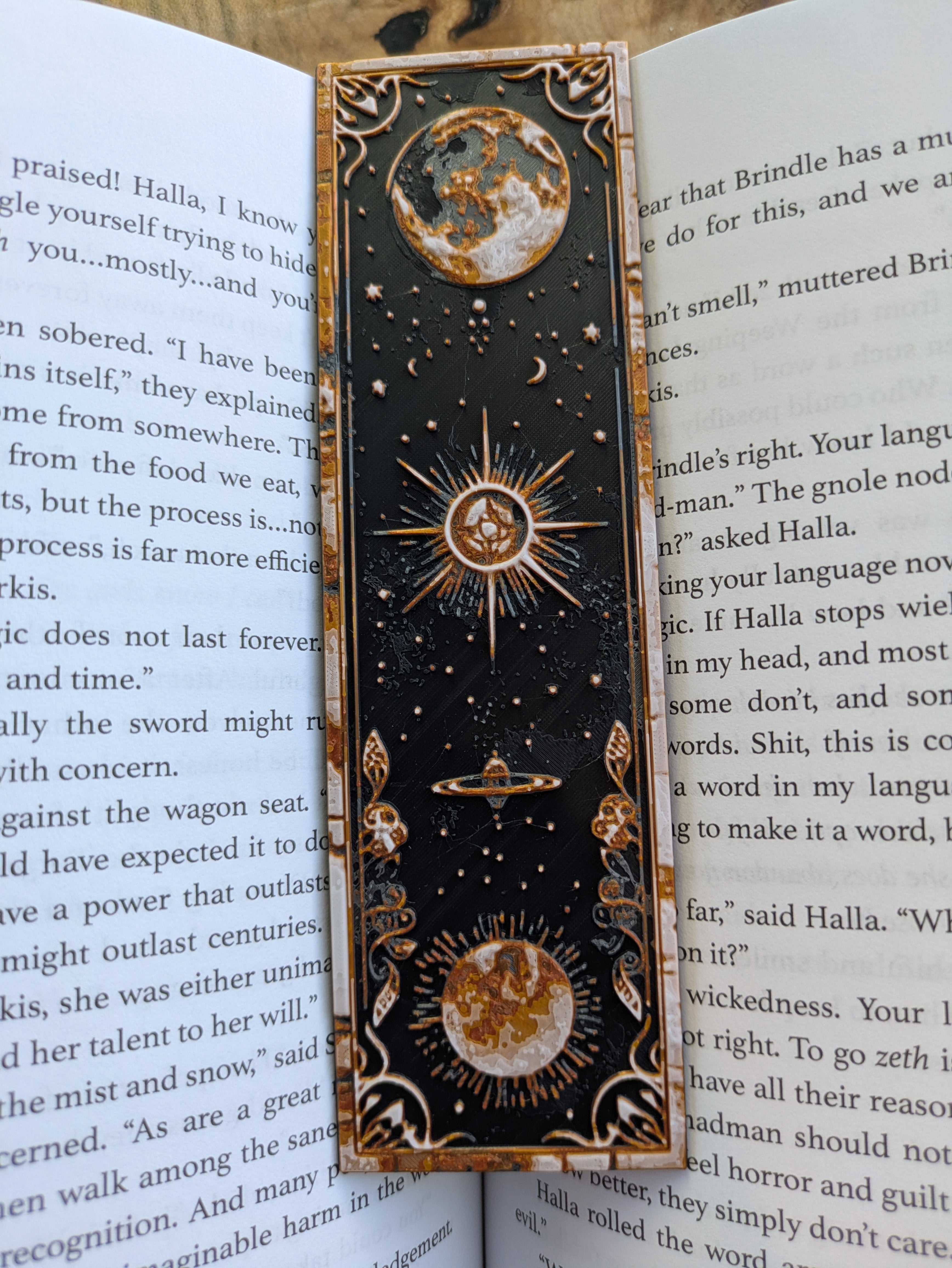 3D-printed bookmark with a celestial Saturn and crescent moon design in gold and white on a black base.