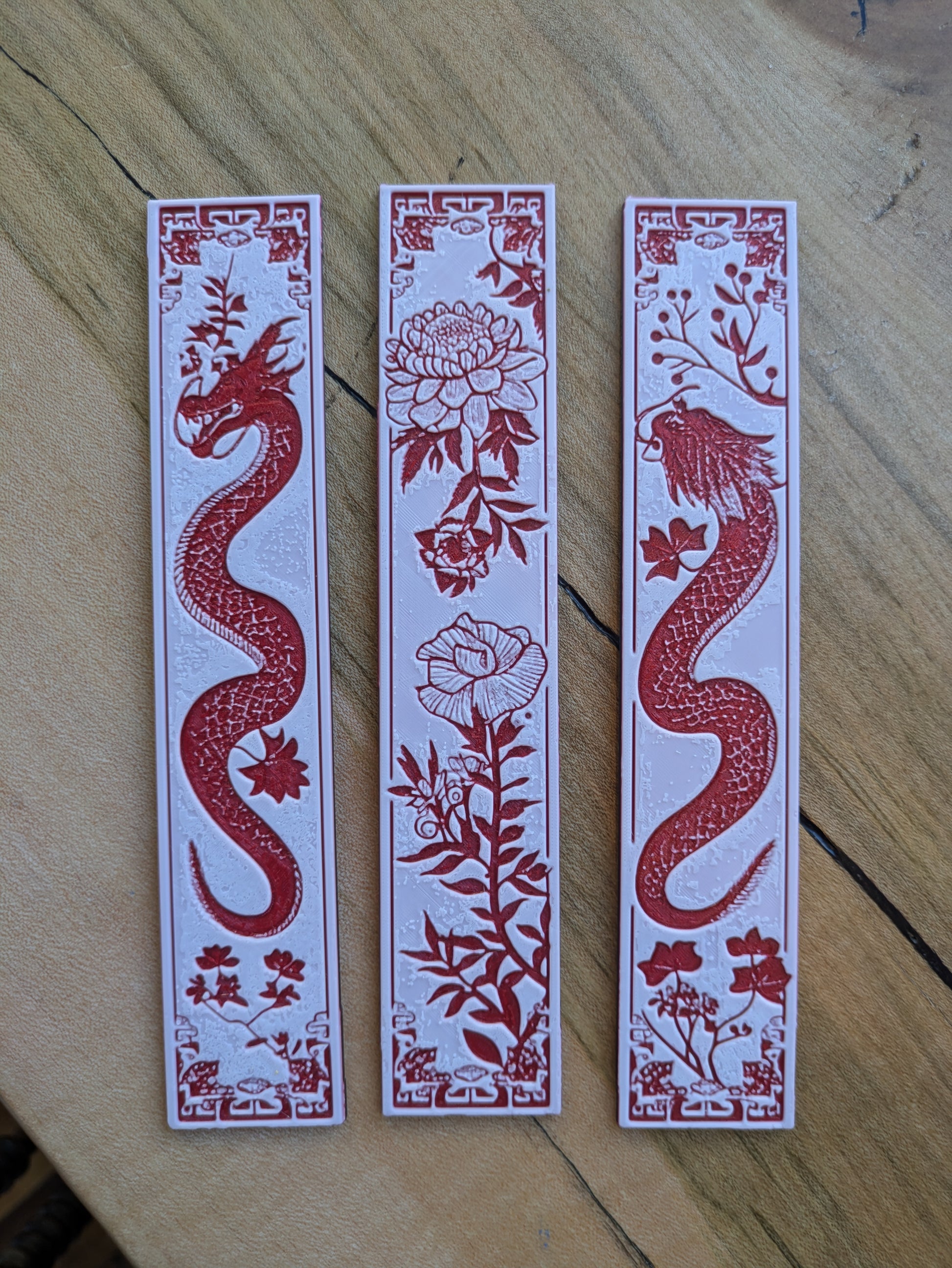 A set of three bookmarks, including
 2 red dragons and  floral blossoms. Displayed artistically on a wooden table.