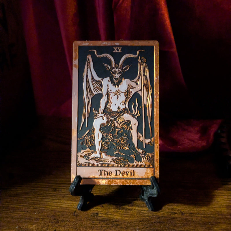 The Devil tarot card bookmark in the Collector Edition, showing the horned, winged figure in copper-toned relief on a black background, displayed on a stand