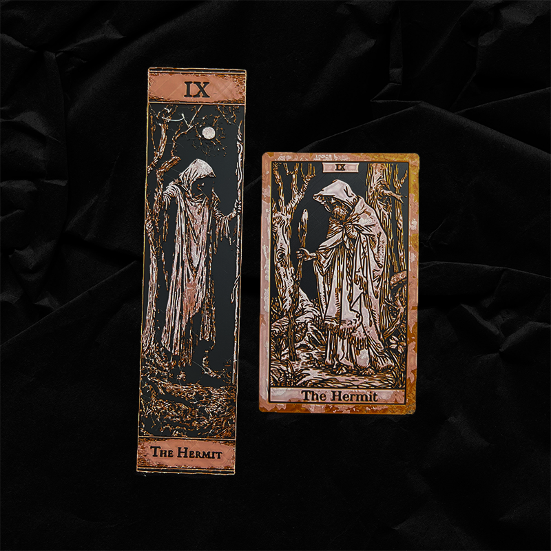 The Hermit tarot bookmark in Classic and Collector Editions side by side on black fabric, showing raised blush and copper relief artwork